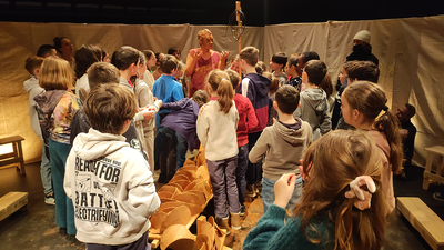 Dans un chapiteau, des enfants sont debout autour d'une comédienne dont le visage est recouvert d'argile.