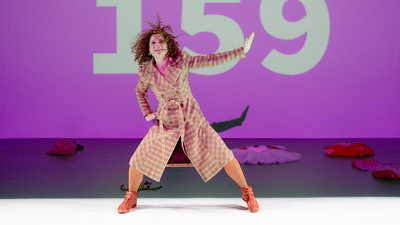 Une danseuse dansant devant un écran violet avec le nombre "159" inscrit dessus.
