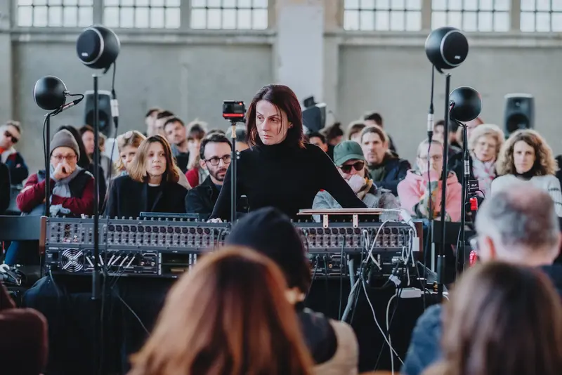 Une femme joue de la musique électronique avec des enceintes, entourée de spectateur·trices assis·es autour d'elle.